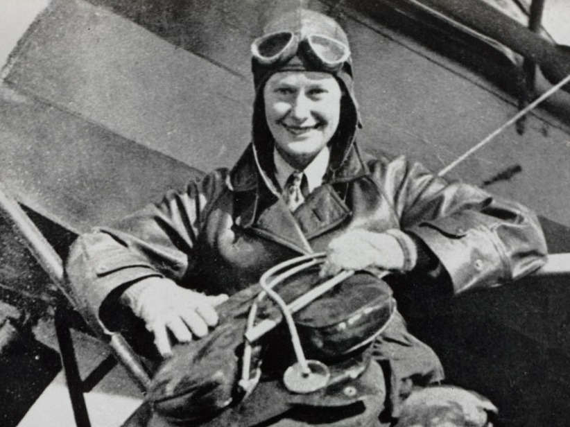 Black-and-white photo showing Nancy Bird Walton standing casually in front of an old-fashioned aircraft wearing full aviation gear, including a cap, with goggles pushed up over her forehead